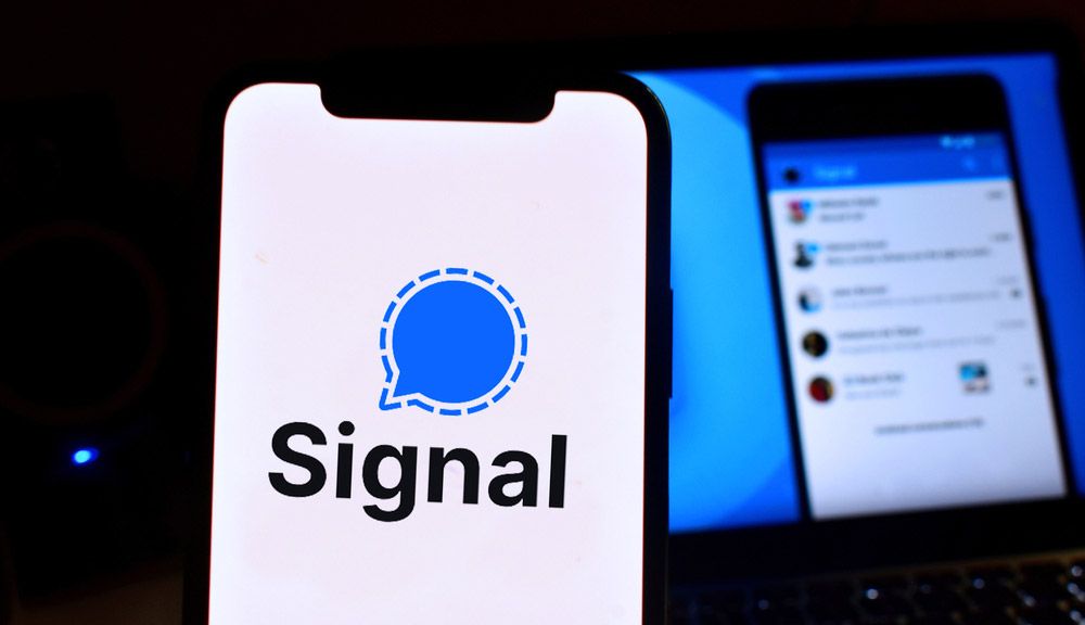 The Signal app logo displayed on an iPhone, with a screenshot of the Signal app in use displayed on a monitor in the background.