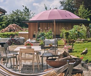 An outdoor scene showing a garden dining set shaded by a pink parasol with firepit and hammock in the foreground