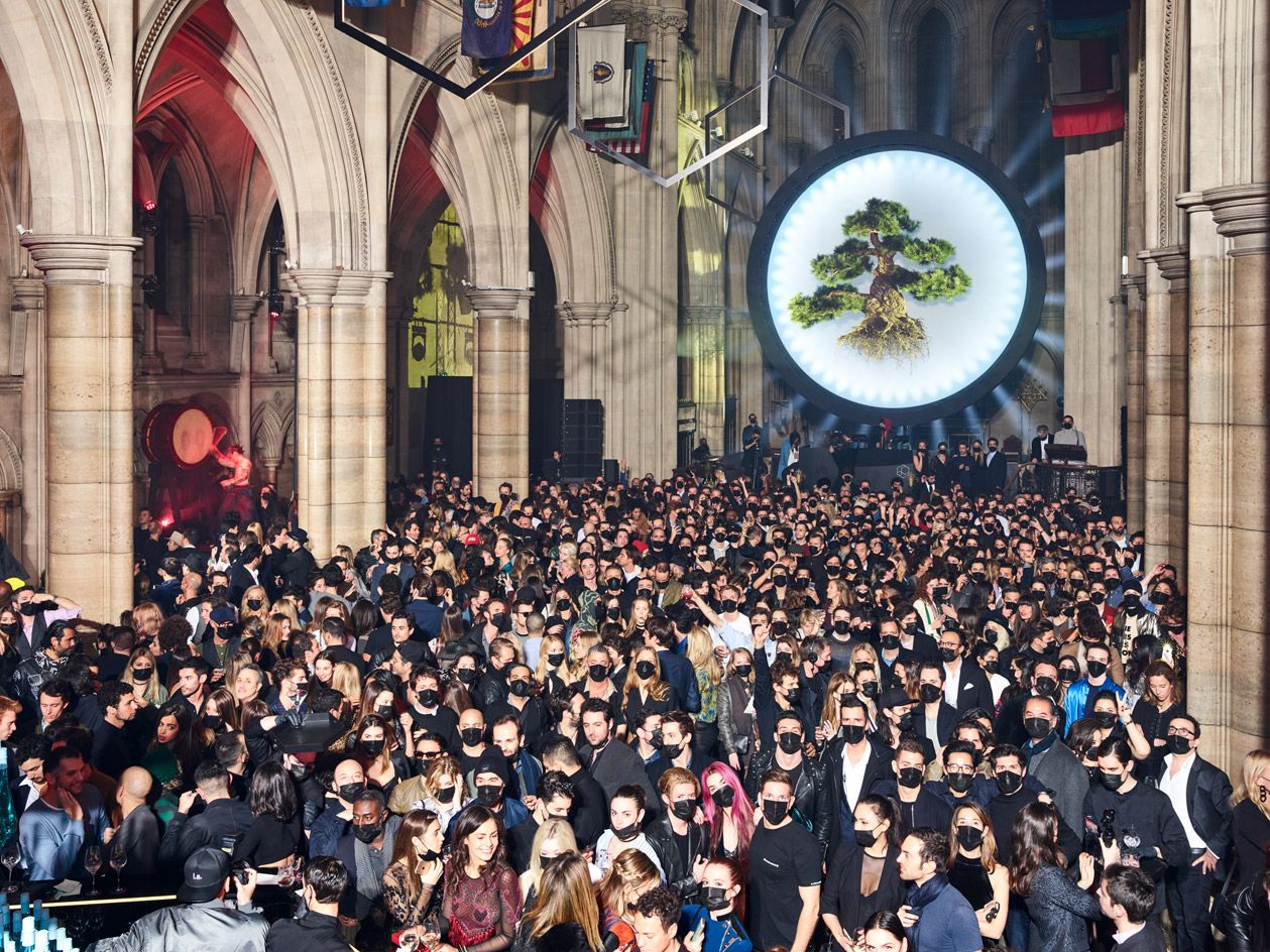 View of a large crowd of people wearing face masks inside The American Cathedral in Paris. There is large round light display with an oversized bonsai tree hanging in front located at the back of the cathedral