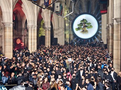 View of a large crowd of people wearing face masks inside The American Cathedral in Paris. There is large round light display with an oversized bonsai tree hanging in front located at the back of the cathedral