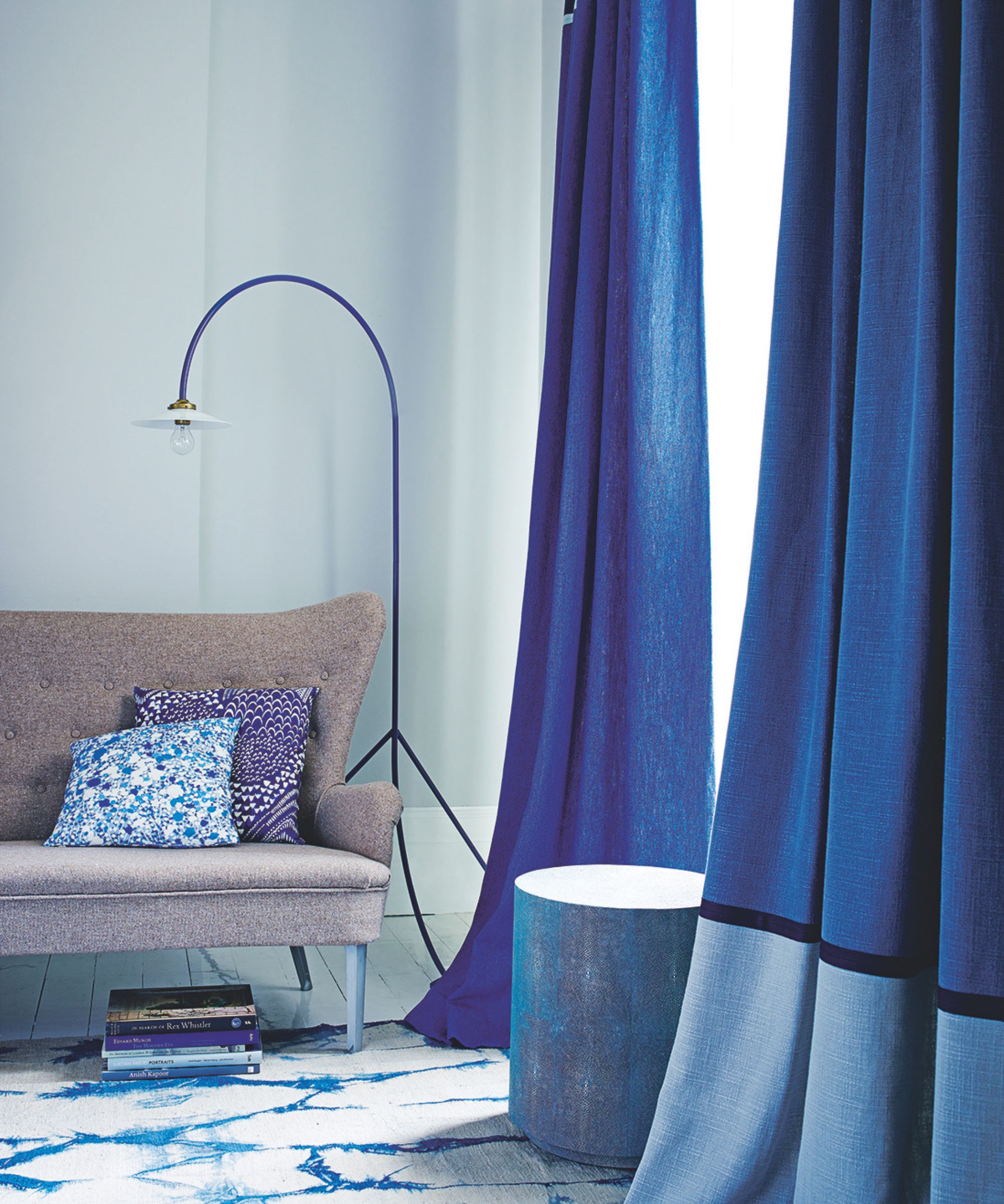 A corner of a pale blue room with blue curtains, a blue and gold stool, brown couch with patterned cushions, a blue curved floor lamp, white and blue patterned rug, and stack of coffee table books.