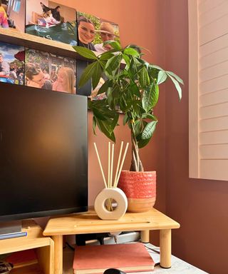 The corner of a home office desk, with a desk monitor riser and a ScentAir reed holder in cream, white design. It looks like a cream donut shape, and there is a potted green plant next to it. Behind there are family photos on terracotta colored walls and a white shutter in view