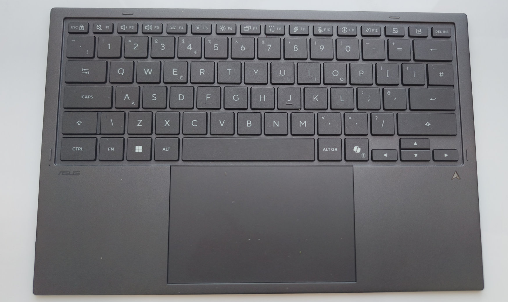 A top-down view of the ASUS Zenbook Duo's detached, dark grey wireless keyboard folio, showing the UK QWERTY layout, function keys, and a large integrated trackpad on the left.