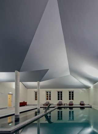 The pool area of the spa. The angular ceiling is gray, and walls with french style windows are painted white. The pool takes up almost the entire room.