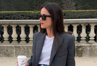 French woman sitting on bench wearing white shirt, jeans, and gray blazer, holding coffee cup, and wearing sunglasses.