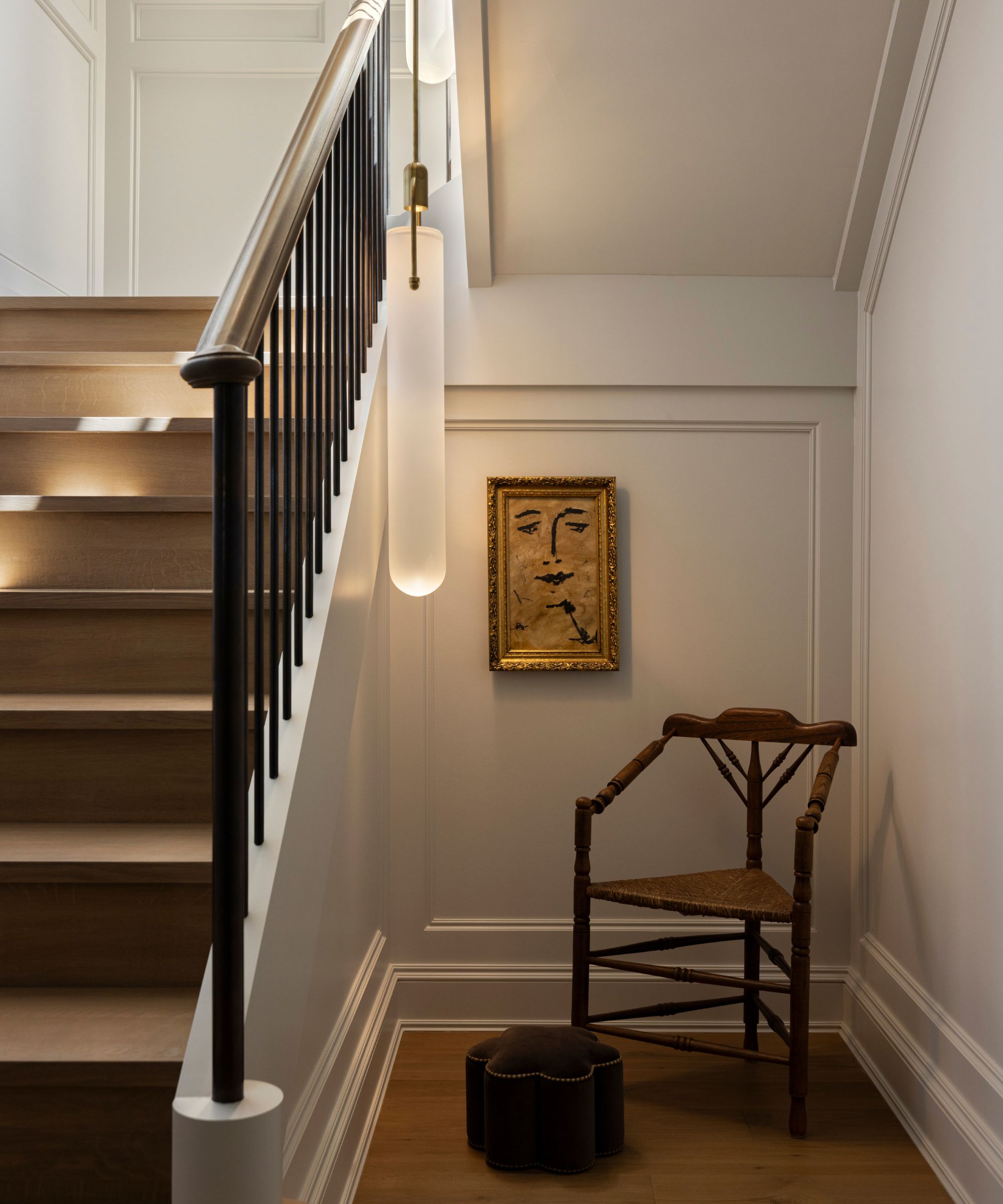 Staircase with iron railing and wooden stairs, cream walls and a little nook with an antique chair, velvet ottoman, and a vintage print framed in a gold frame on the wall