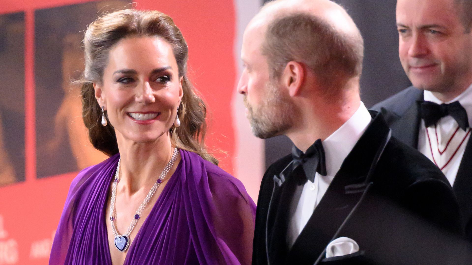 Kate Middleton arrives at the "Digger" Premiere wearing a royal purple long-sleeve maxi dress with a long necklace and drop earrings. She walks alongside Prince William.