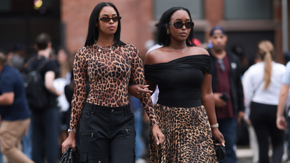  A guest (L) wears Miu Miu brown sunglasses, gold pendant chain necklace, brown animal print / leopard print sheer long sleeve top, black cargo long pants with pockets and Bottega Veneta black woven leather pouch / clutch bag; Next to her a guest (R) seen wearing Celine brown oval Triomphe sunglasses, Dior vintage gold earrings, diamond pendant necklace, black cotton off-shoulder top, brown animal print / leopard print flared pleated long skirt and Bottega Veneta black woven leather mini Jodie bag, outside the Alice + Olivia by Stacey Bendet Spring 2025 presentation, during the New York Fashion Week, on September 07, 2024 in New York City. 