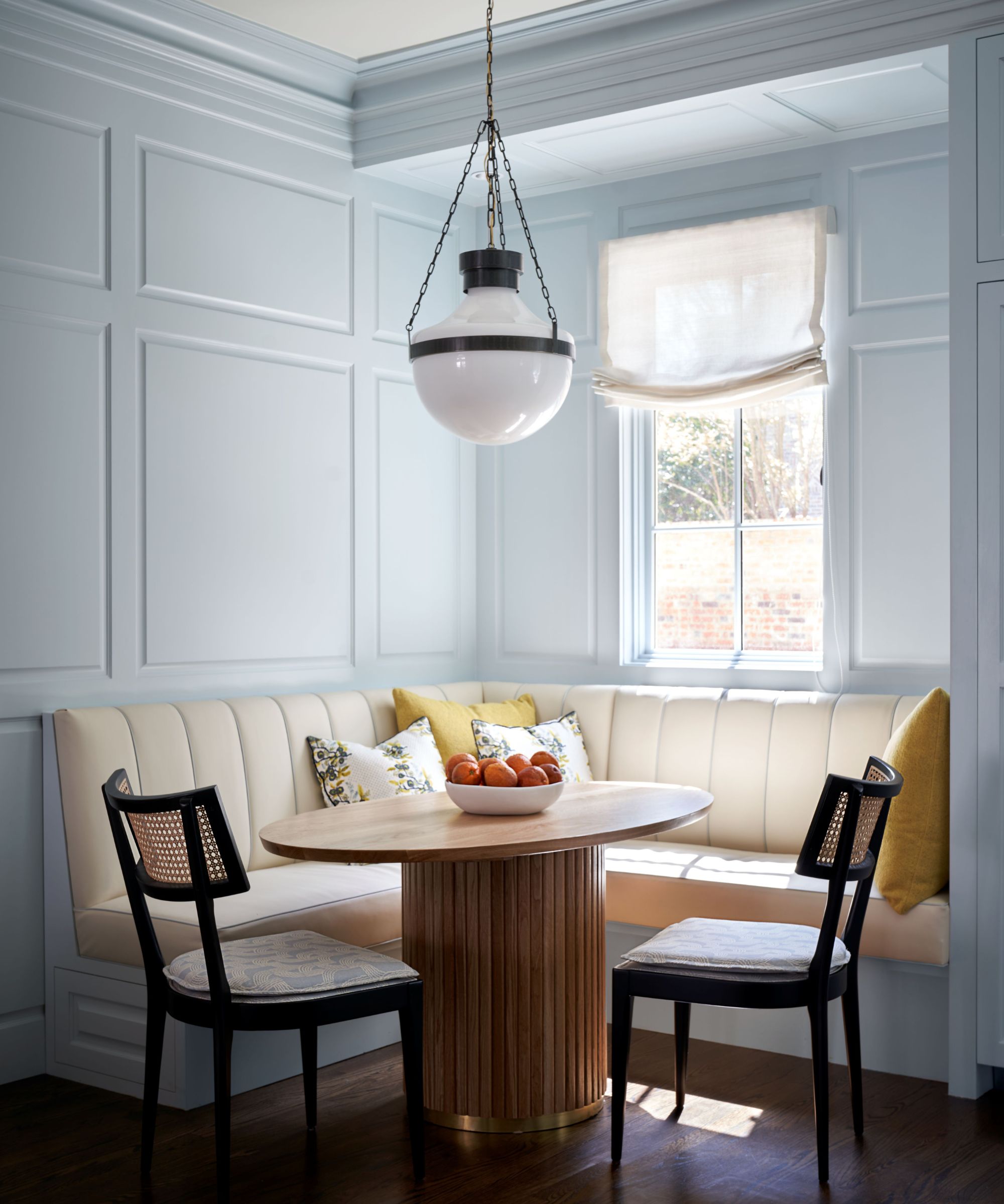 Dining nook with built-in cream banquette seating, walls painted in Farrow &amp;amp; Ball's Borrowed Light, a low glass lantern pendant, a round wooden table, and two black wicker dining chairs.