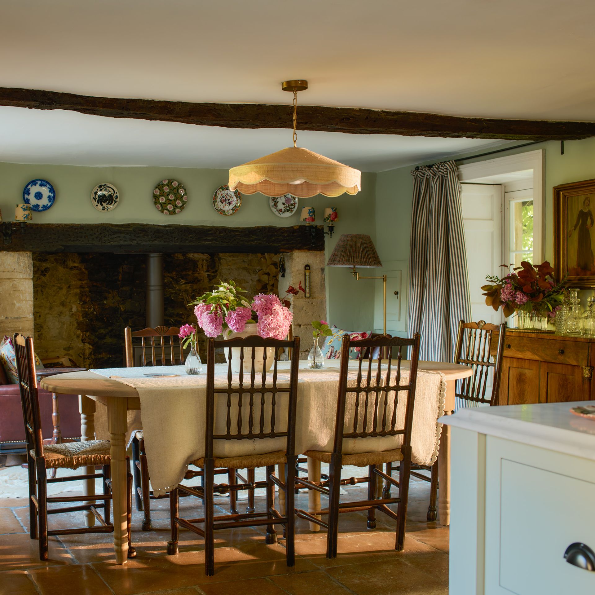 extendable dining table at the end of an open plan kitchen wtih sage walls rattan lampshade above table and inglenook fireplace behind