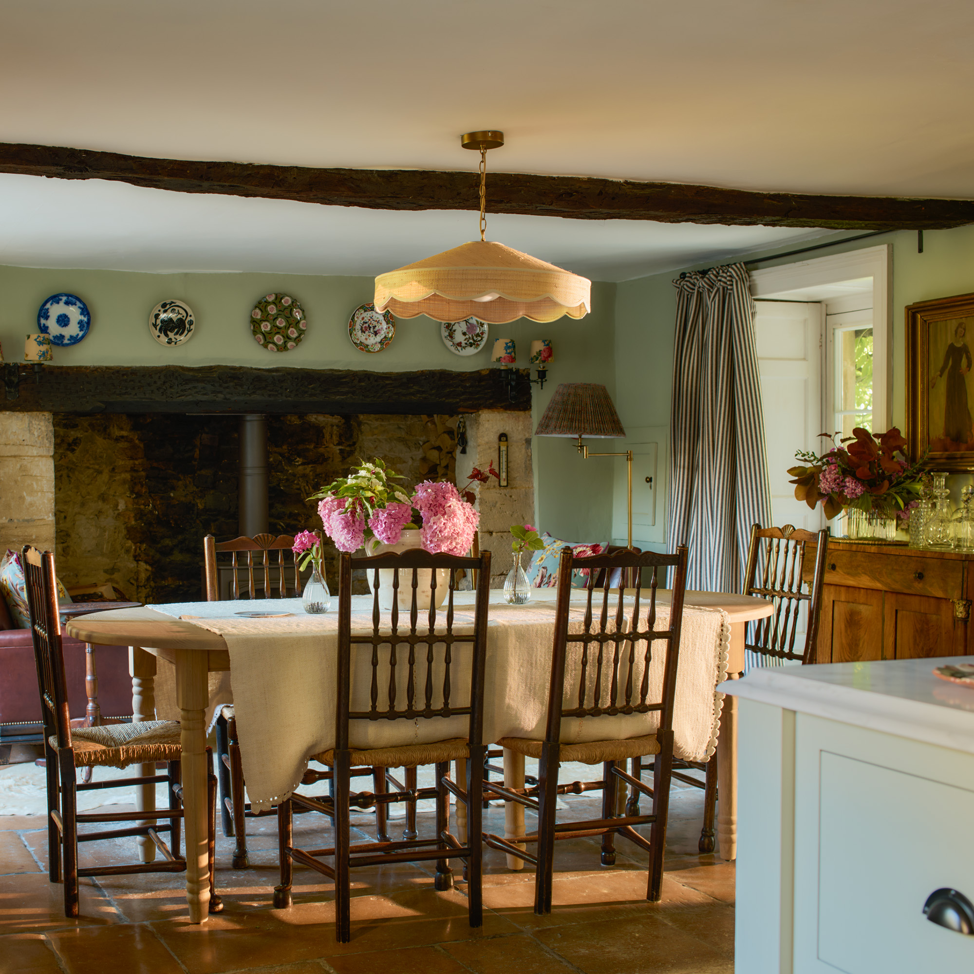extendable dining table at the end of an open plan kitchen wtih sage walls rattan lampshade above table and inglenook fireplace behind