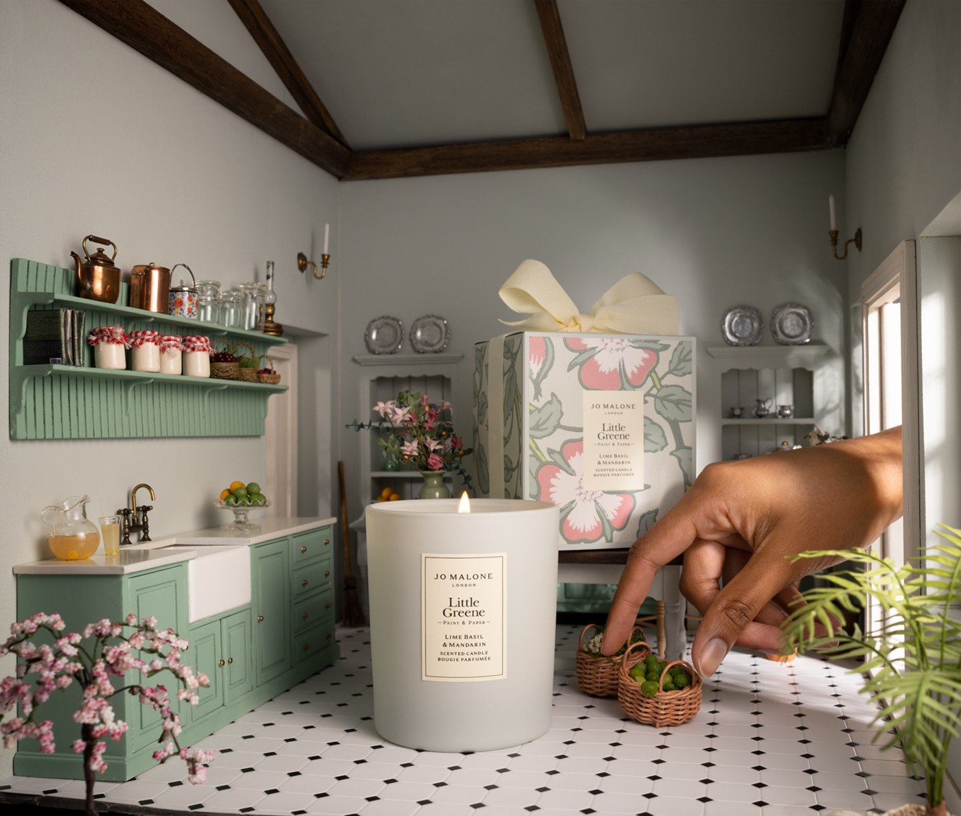 Image of a miniature kitchen that is painted in a pale sage green color with darker sage green cabinetry and shelving. There is a hand reaching in from the side, touching miniature wicker baskets. There is a regular-sized candle that's in the middle of the room.