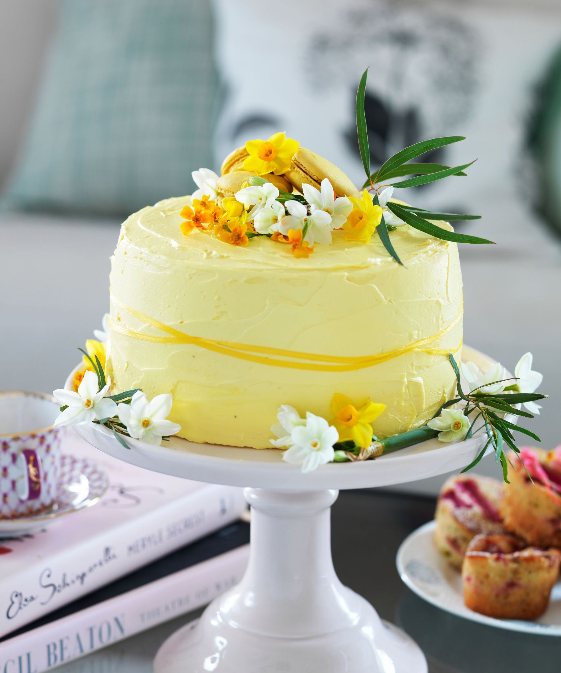 Yellow iced cake decorated with flowers and macarons on a white ceramic cake stand