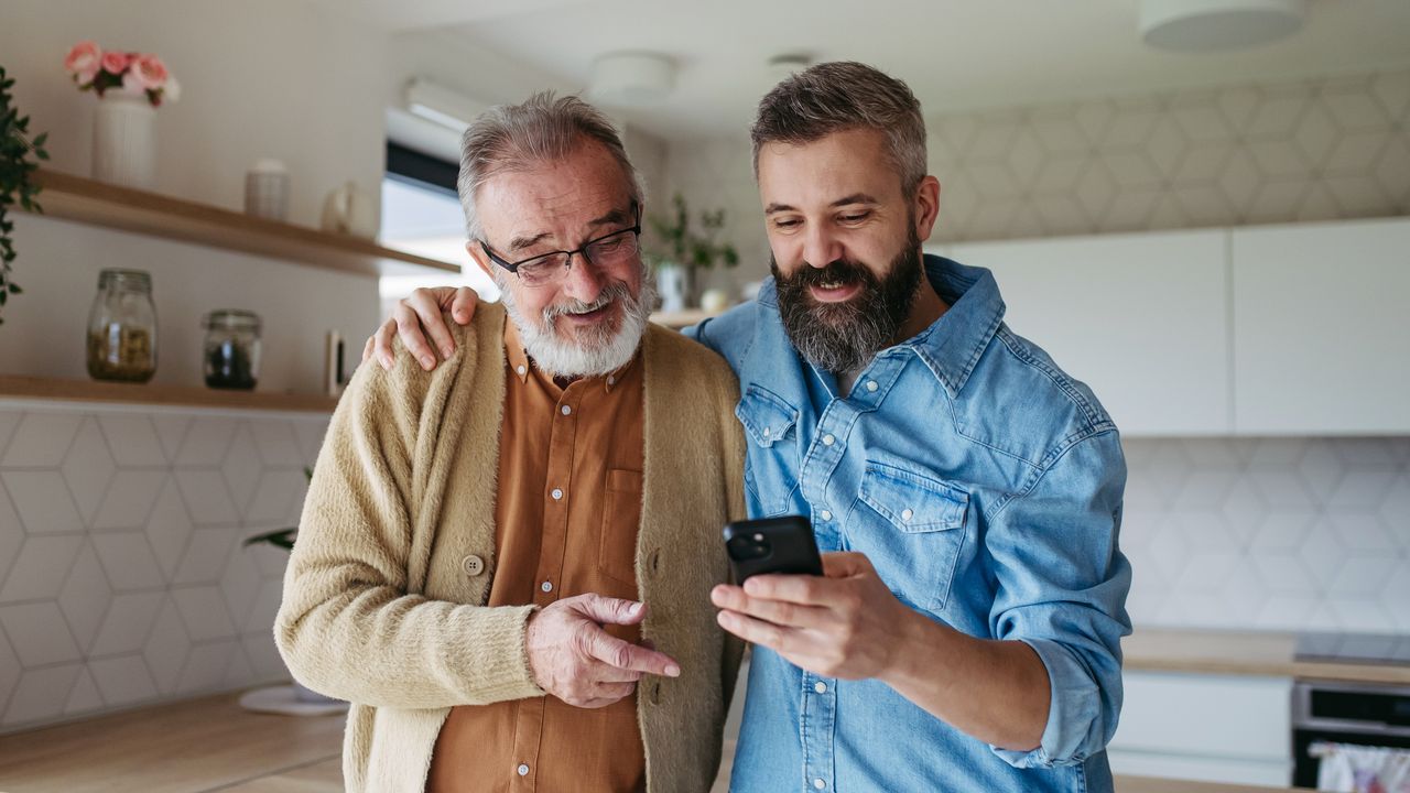 A dad and his adult son stand next to each other and look at the son&#039;s smartphone.