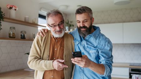 A dad and his adult son stand next to each other and look at the son's smartphone.