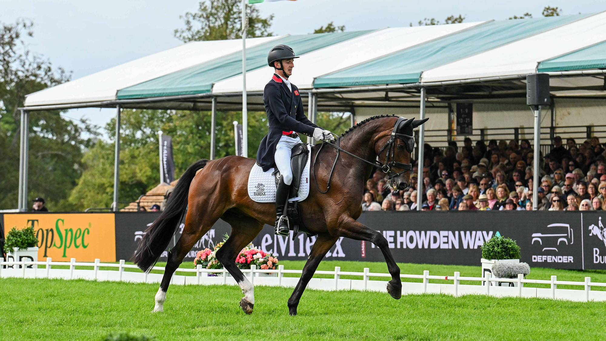Tom Jackson, a rider in a black tailcoat with red trim, rides bay horse United 36 in the Burghley grass dressage arena