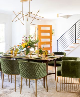 A bright white dining area with a wooden dining table with dark green plates and velvet chairs around it, yellow glasses, a vase of purple, yellow, and orange flowers, and a brushed gold abstract chandelier above it
