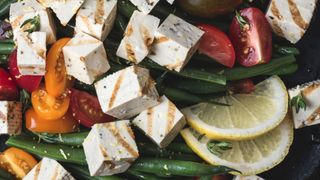 Cubes of tofu drizzled with sauce, on a bed of green vegetables, lemons and tomatoes