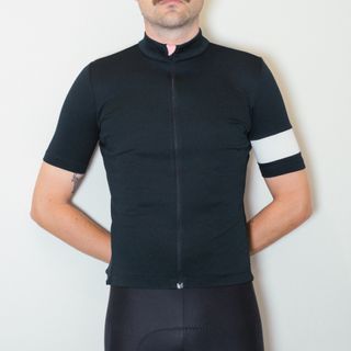 A white man in a black cycling jersey and black cycling shorts stands against a white wall