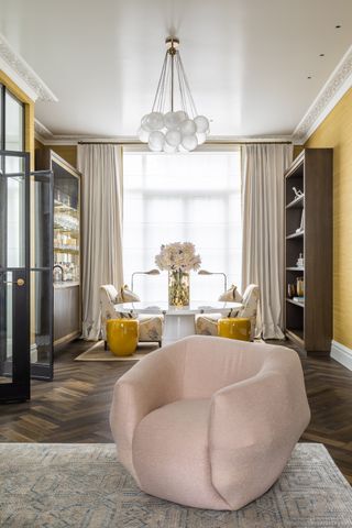 yellow living room with dark wood herringbone floor, rug, pink armchair, yellow stools, patterned armchairs, round white table, bookshelves, globe ball pendant