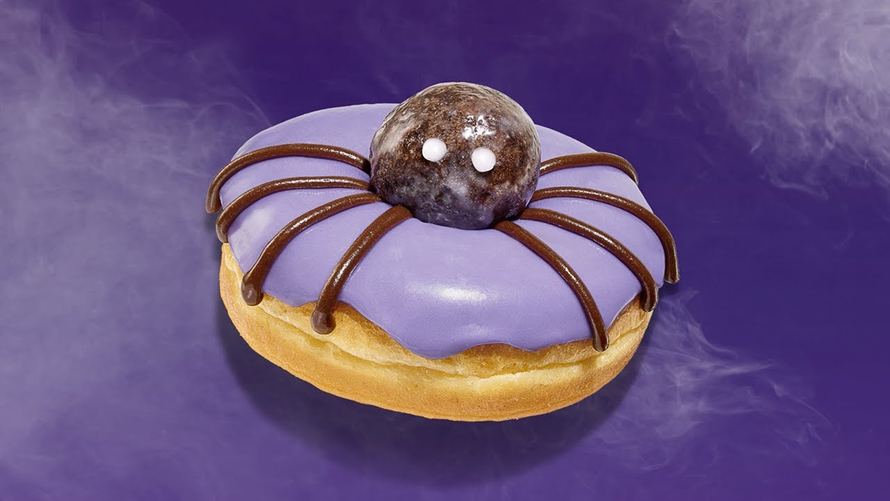 A Halloween-themed donut with purple frosting, chocolate drizzle legs, and a glazed chocolate munchkin in the center decorated with white eyes to look like a spider.