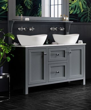 A sophisticated bathroom featuring a dark grey double vanity unit with shaker-style doors and polished chrome knobs. Two white countertop basins with elegant curves sit atop a marble-effect worktop. Above, a large wall mirror with a built-in grey shelf reflects light, complementing the dark tones.