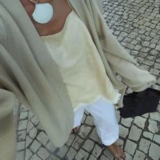 Image of @anoukyve wearing a neutral transitional outfit: white jeans, cream slip top, cardigan and shell necklace.