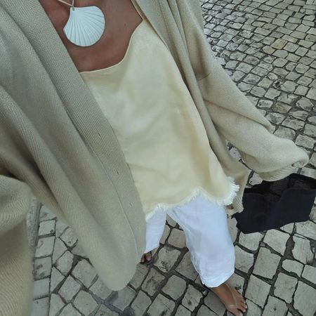 Image of @anoukyve wearing a neutral transitional outfit: white jeans, cream slip top, cardigan and shell necklace.