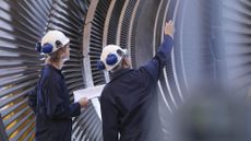 A pair of engineers examine wearing white hard hats stood in front of a large turbine engine