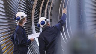 A pair of engineers examine wearing white hard hats stood in front of a large turbine engine