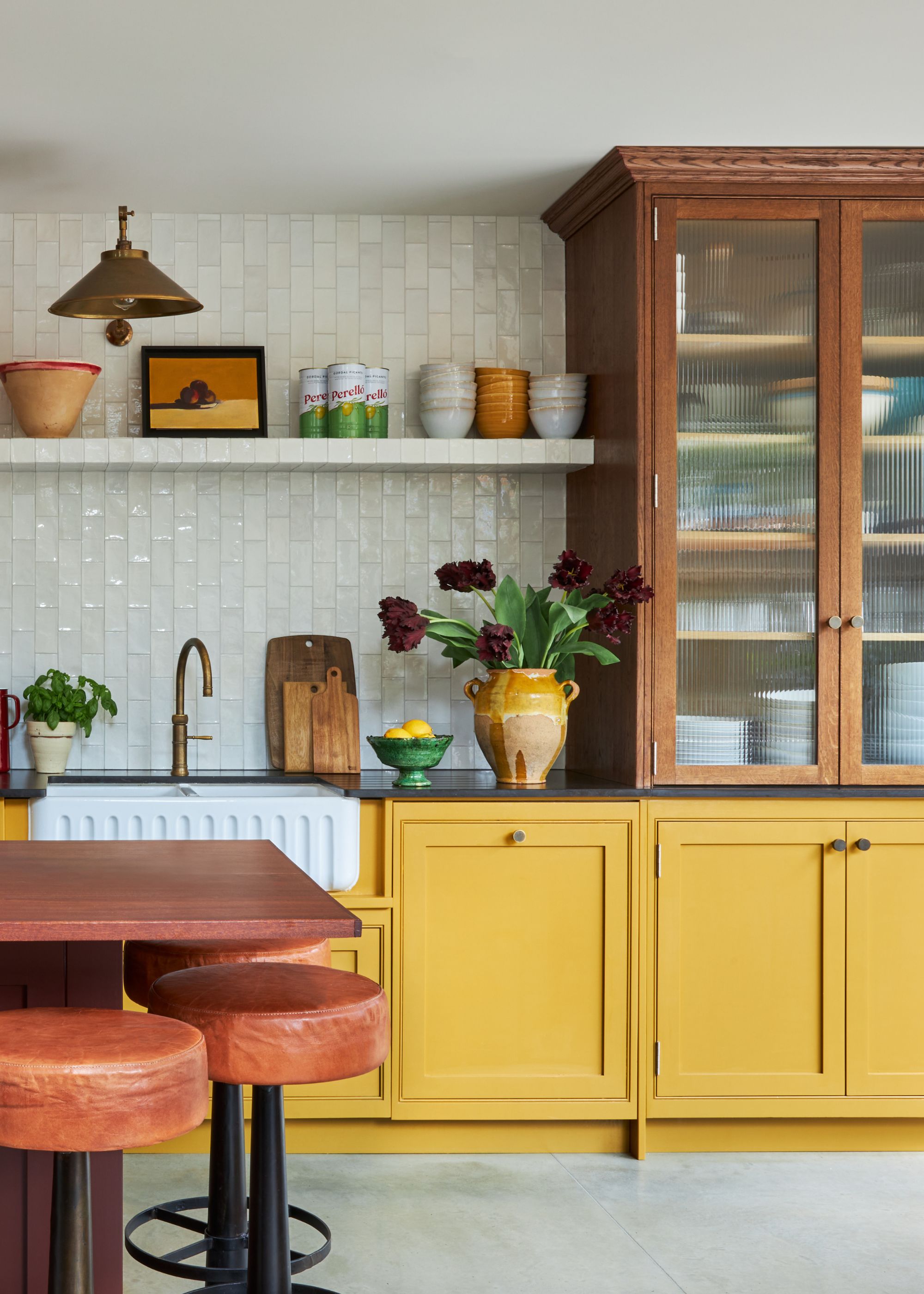 A kitchen with dark yellow lower cabinets, concrete flooring, warm white zellige tiles on the wall, a wooden pantry unit, and modern rustic decor.
