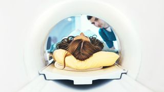 female patient with long hair lying down in an MRI; we can see the top of her head and a medical provider peering in the machine on the other side