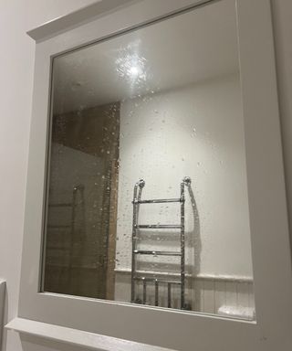 A wet mirror with a white wooden border and silver towel rail in the reflection