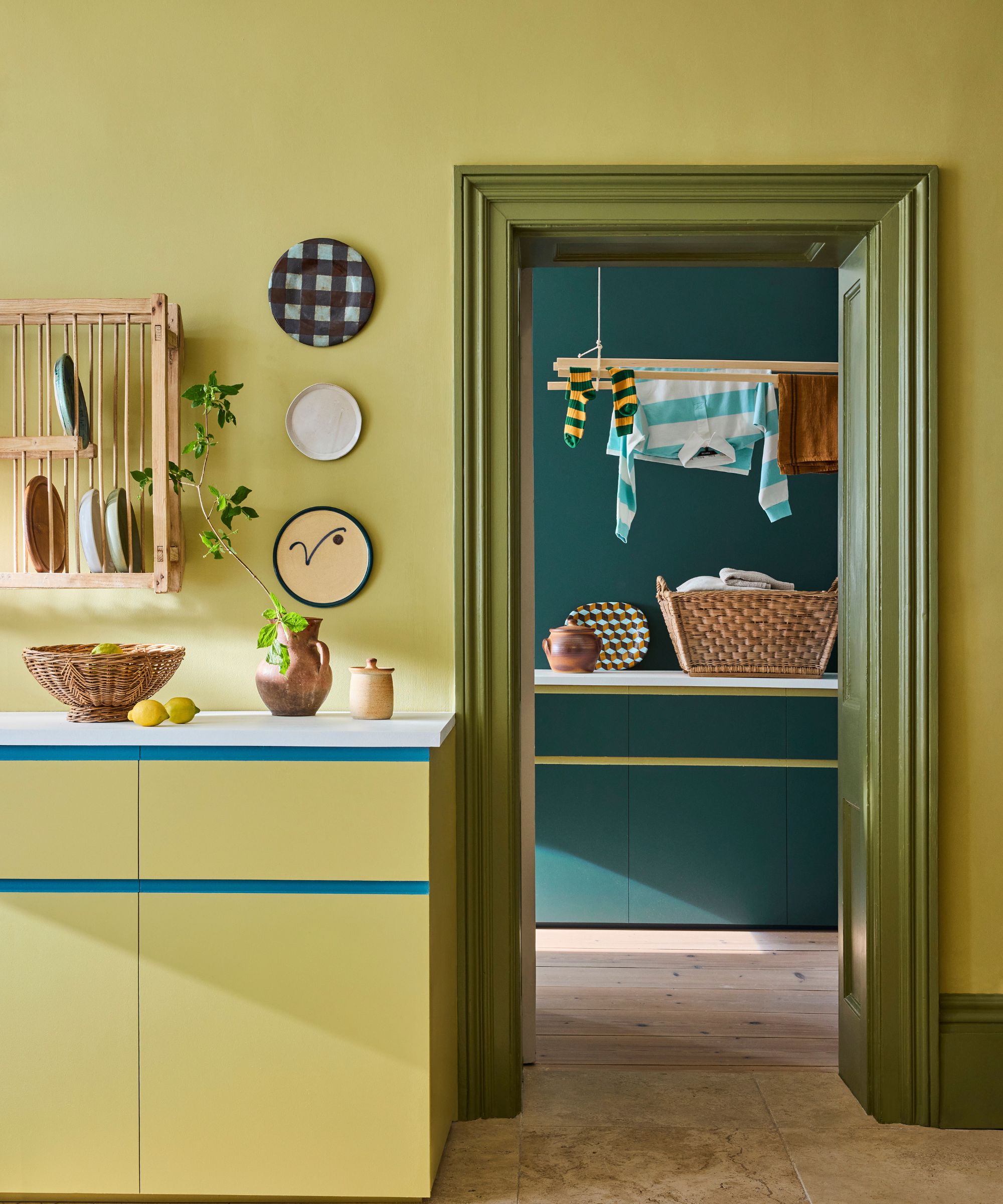 Citron green kitchen with plates hanging on the wall, a dark green door frame, looking into a teal laundry room with clothes hanging up.