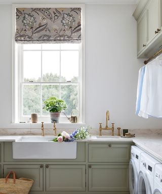 Muted sage green cabinets in laundry room with double utility sink, brass taps, white walls, and floral window blind