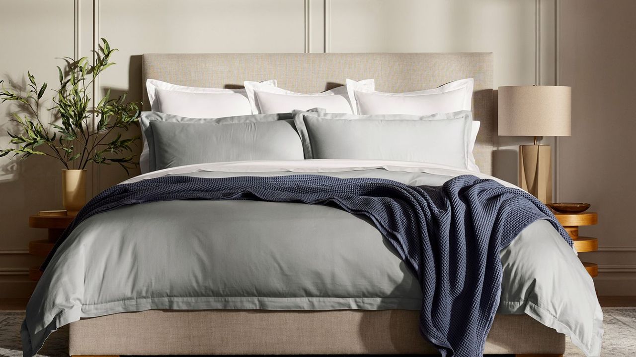 Grey and white bedding with a navy blanket on a bed against a beige wall.