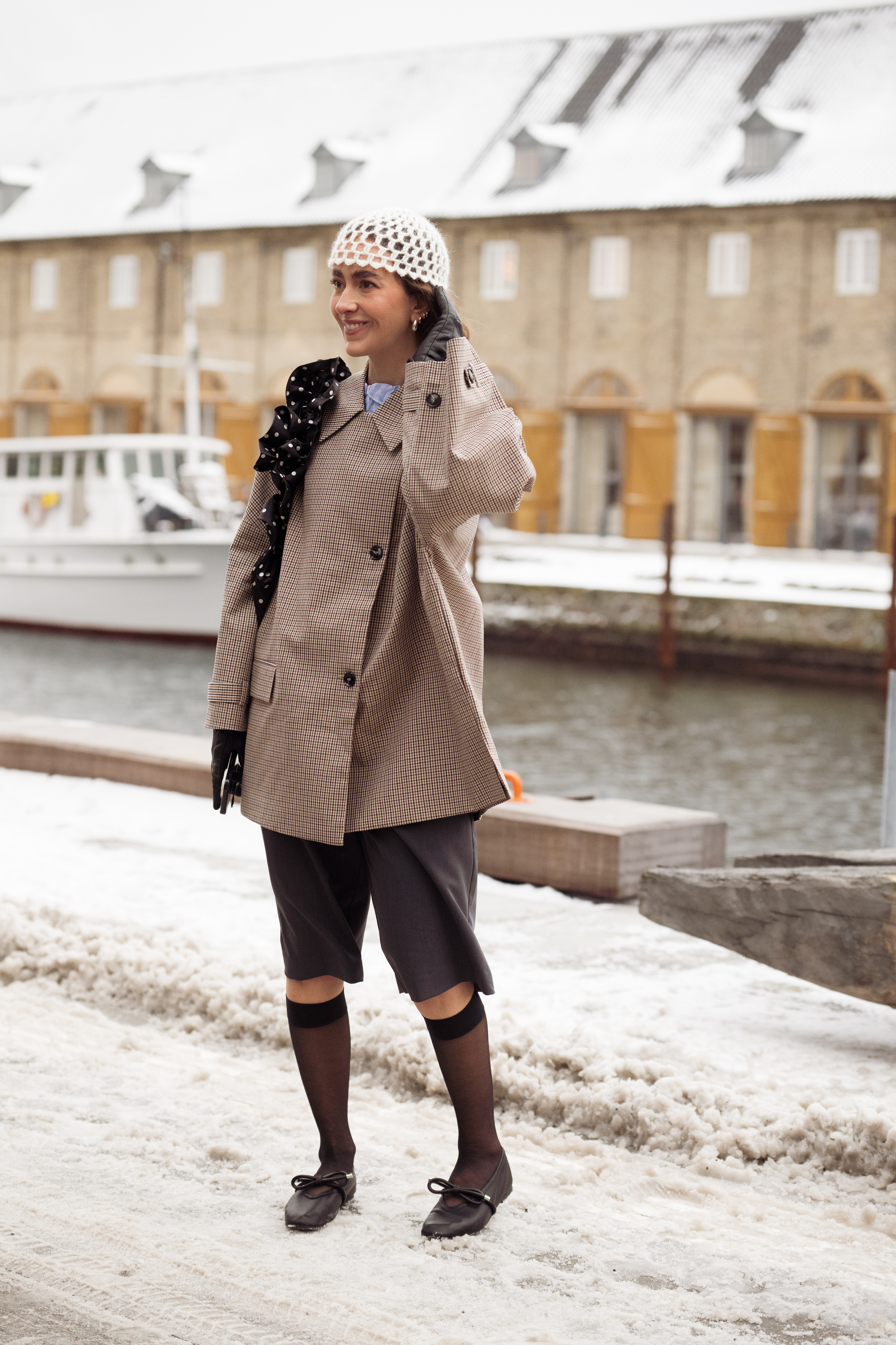 COPENHAGEN, DENMARK - JANUARY 28: A guest wears a beige and brown houndstooth patterned trench coat, a white crocheted net hat, dark knee-length tailored shorts, sheer black knee-high socks, black leather ballet flats with bow details, black leather gloves, and a black polka-dot ruffled scarf draped over one shoulder outside the Herskind show during Copenhagen Fashion Week AW26 on January 28, 2026 in Copenhagen, Denmark. (Photo by Raimonda Kulikauskiene/WireImage)