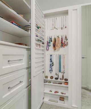 An open door in a wall with colorful necklaces hung up in the wall and on the door, three shelves (two with bracelets and one with boxes) and wall shelves and drawers to the left
