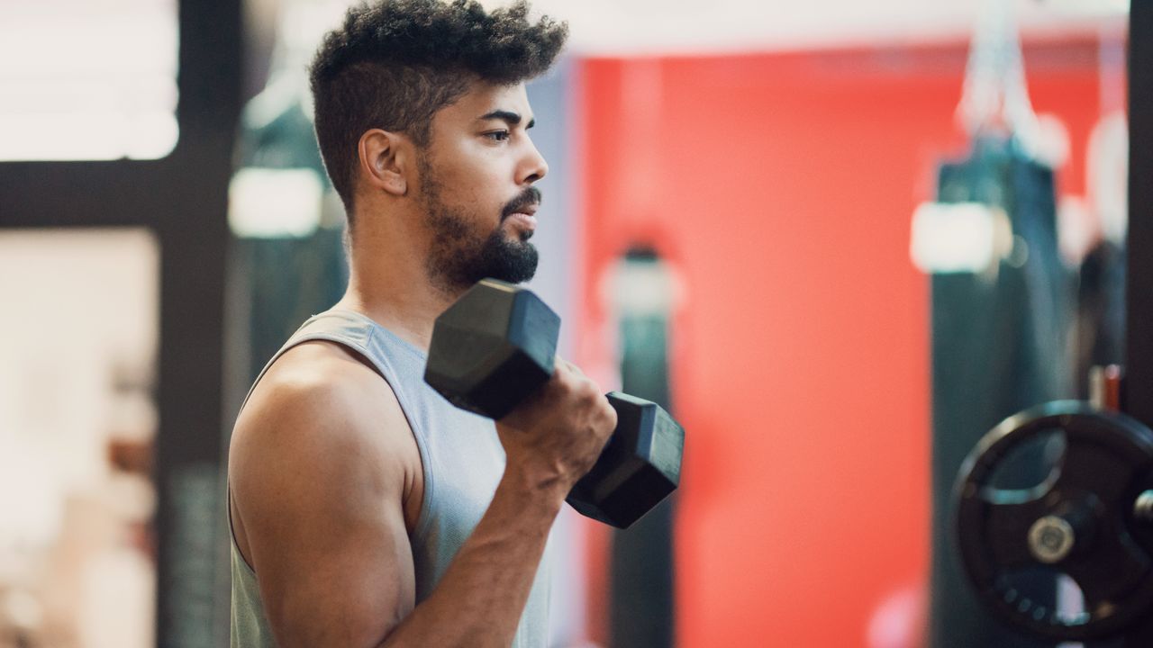 A man performs biceps curl in a gym. He has a heavy dumbbell in his right hand, and his elbow is bent so that the dumbbell is near his shoulder.