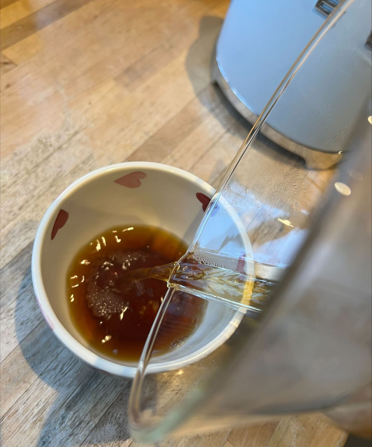 Pouring coffee into a white teacup with pink flowers on a wooden countertop with the Chemex coffeemaker. In the background is a baby blue Smeg kettle.