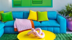 Grey living room with bright blue sofa, green rug and artwork, yellow coffee table and multicolor cushions