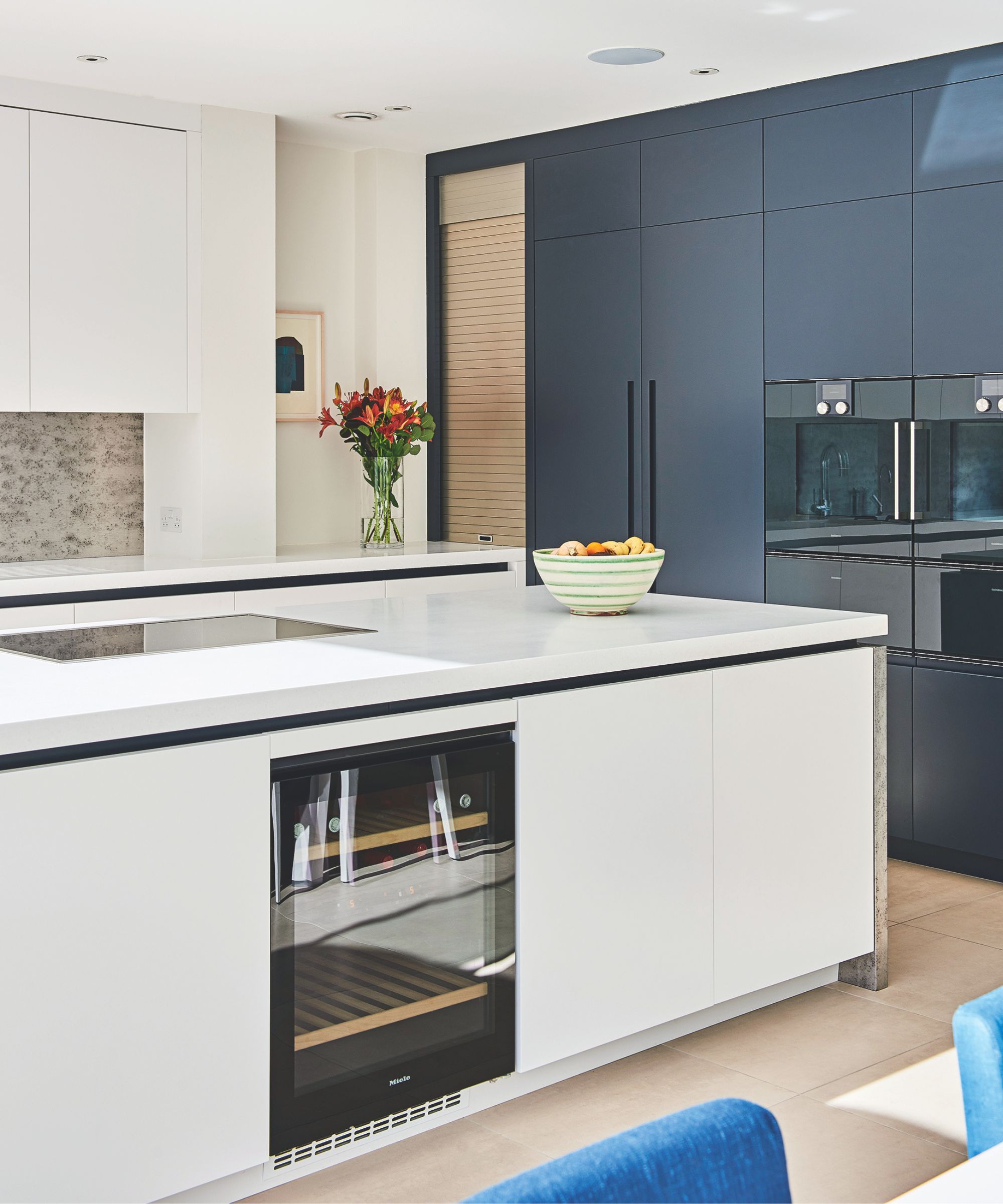 A tidy white kitchen with blue cabinets, a vase of red and orange flowers, and a white and green striped fruit bowl. The flooring is light and wooden. In the foreground are the backs of two blue velvet chairs.