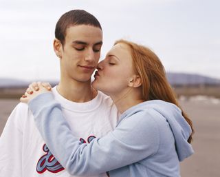 A teenage girl with her arms around a teenage boy, leaning in to kiss him on the cheek