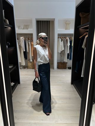 Eliza Huber in the dressing room at Banana Republic wearing a white sleeveless T-shirt with wide-leg dark-wash denim, a headscarf, a black mini bag, and black flat slides.