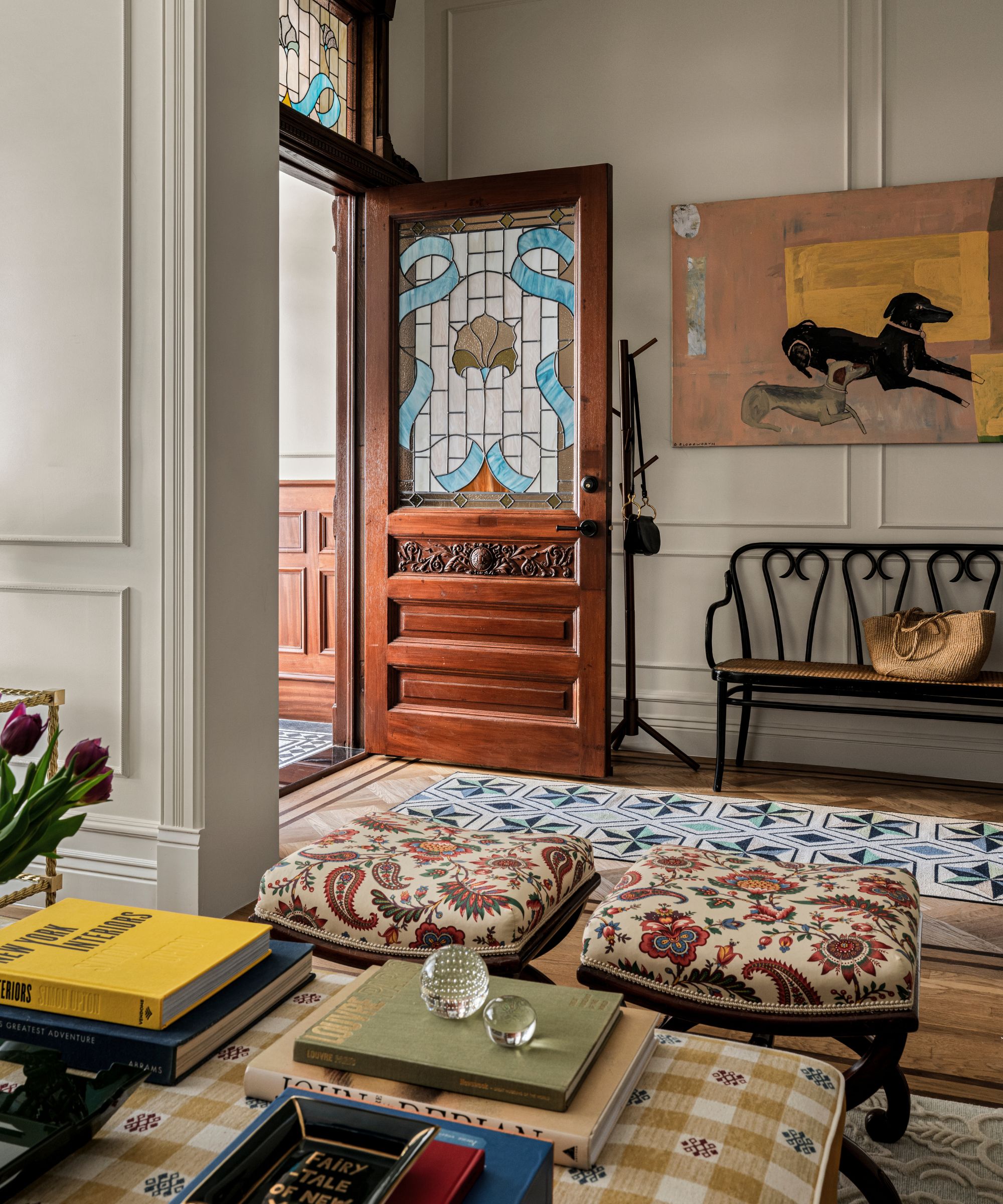 a new york brownstone entryway with mahogany front door with stained glass panels leading into an open plan entry with a bench, artwork of dogs, and a small sitting area with patterned ottomans
