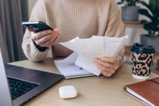 Close-up of a woman reviewing receipts while holding a smartphone beside an open laptop at a cozy desk. 