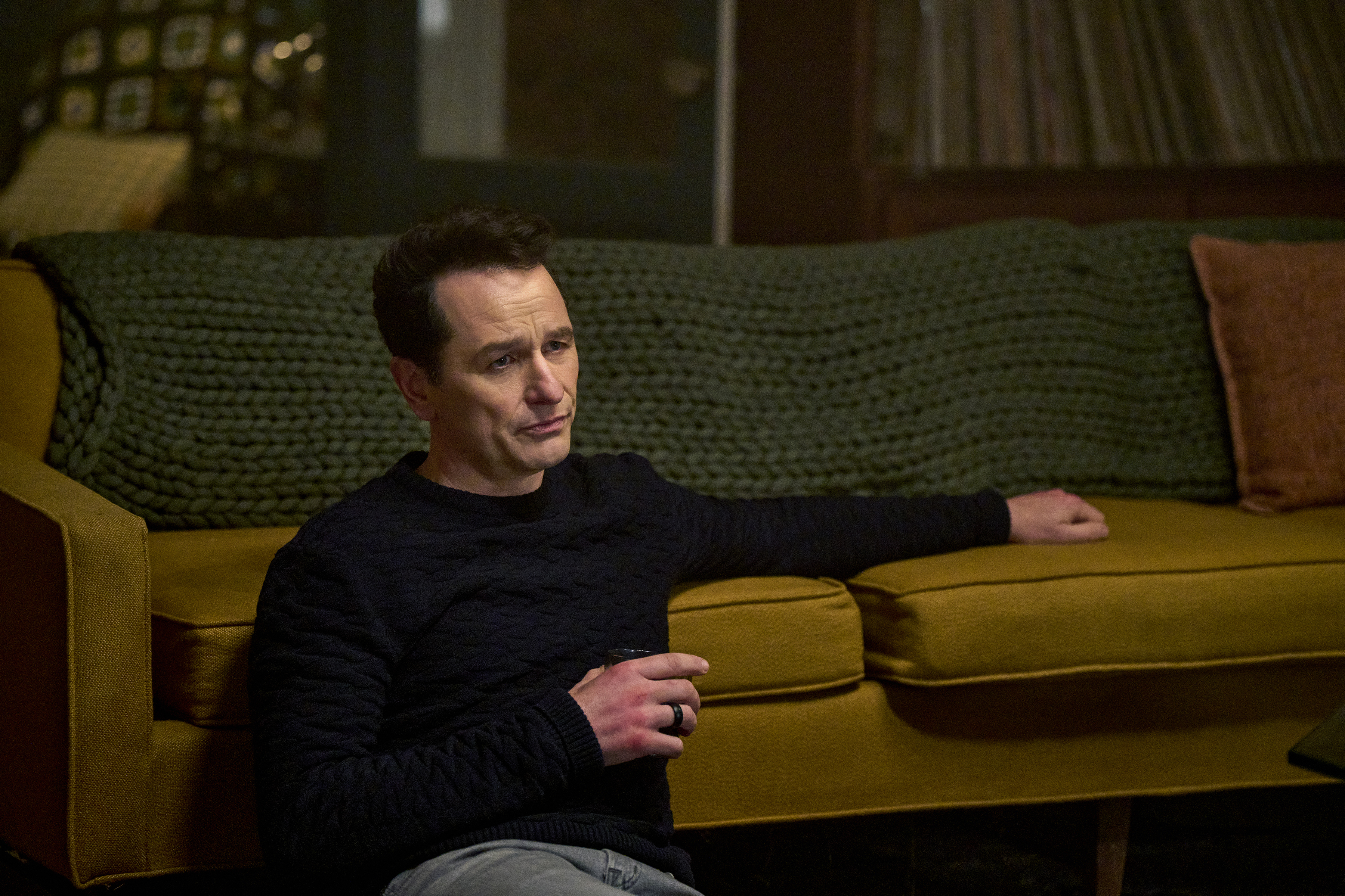 matthew rhys as Nile Jarvis sitting on the floor in front of a sofa in the beast in me