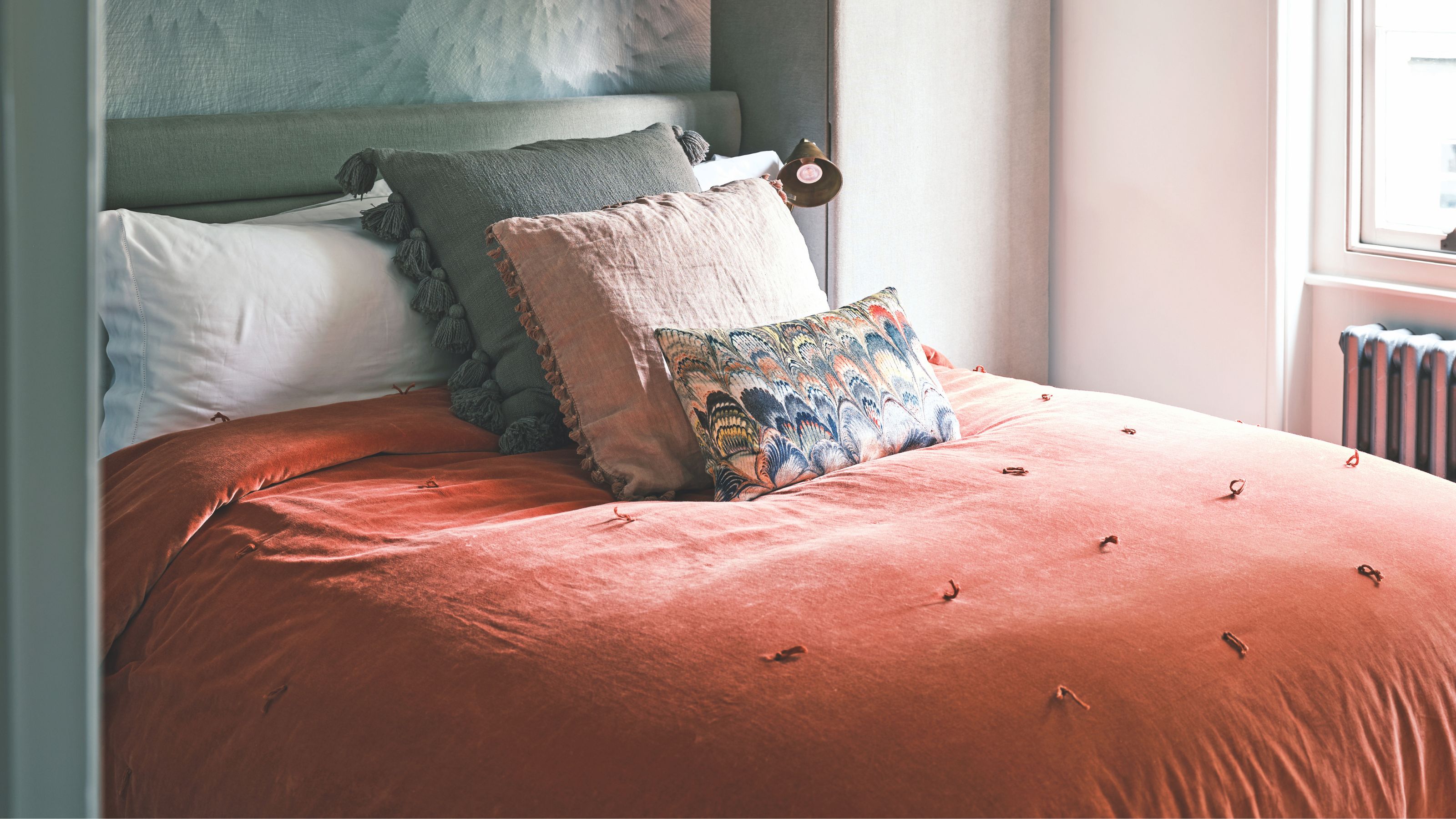 Double bedroom with burnt orange bedding, assorted cushions, white walls, and window with a silver radiator beneath.
