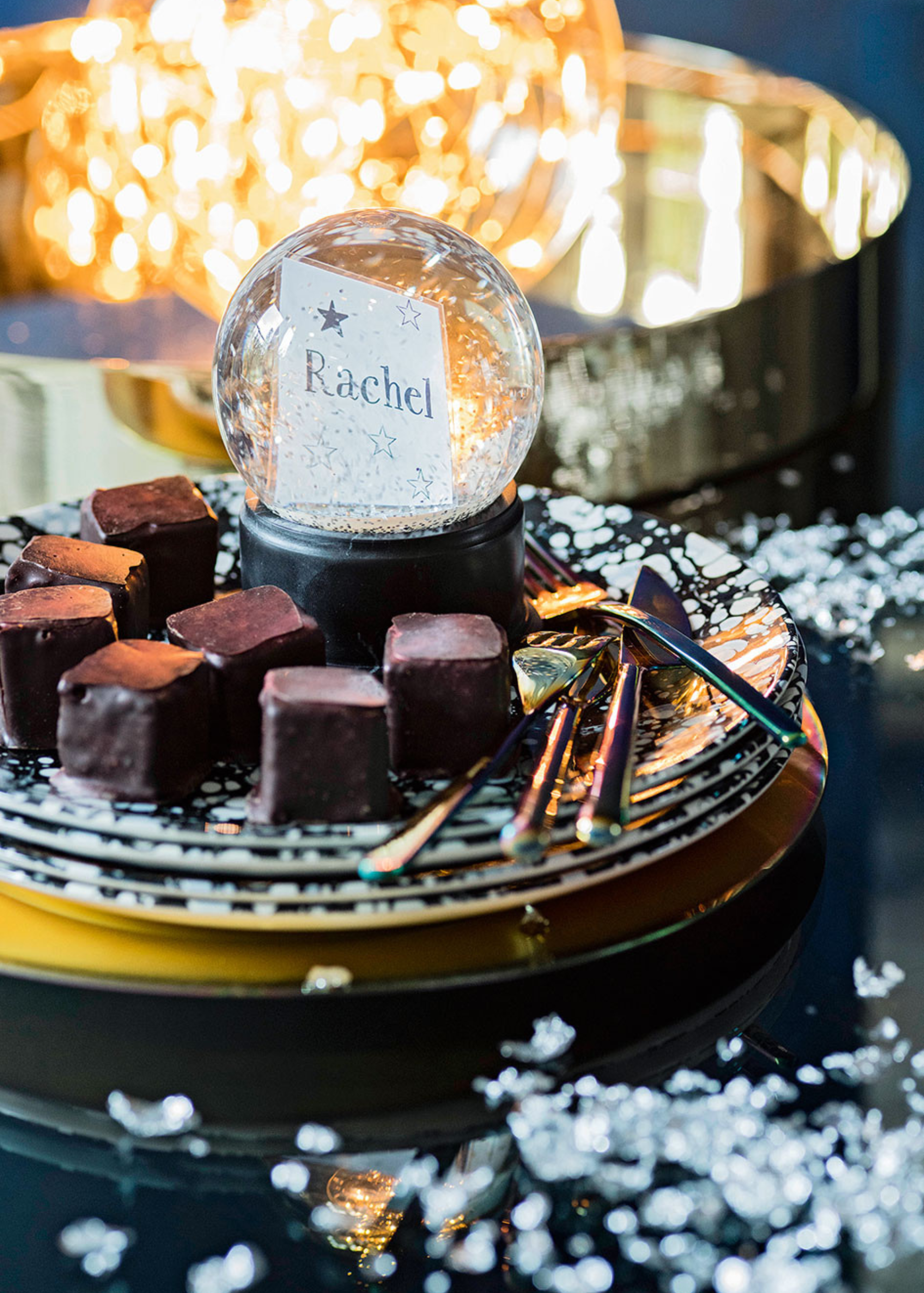 A Christmas table with a pile of plates with chocolates on top and a snow globe with a place setting for &#039;rachel&#039; inside with a lit ball in a gold tray in the background and artificial snow all around