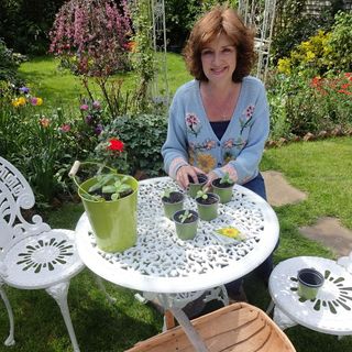 A lady in a blue cardigan tending to potted plants in a garden seated at a white garden table